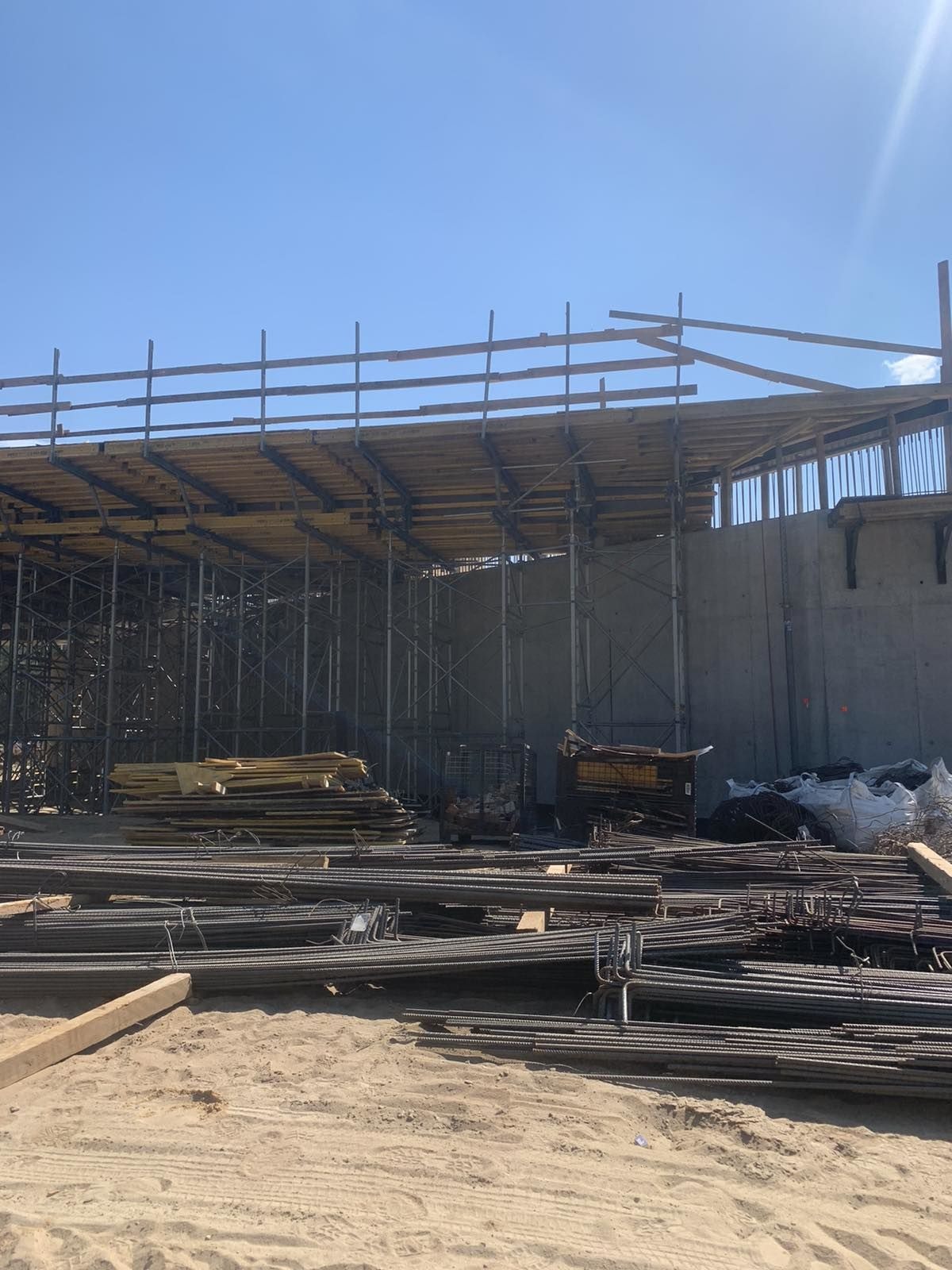 Widok placu budowy w Warszawie z przygotowanymi zbrojeniami, deskowaniem i rusztowaniami na tle betonowej konstrukcji w słoneczny dzień.