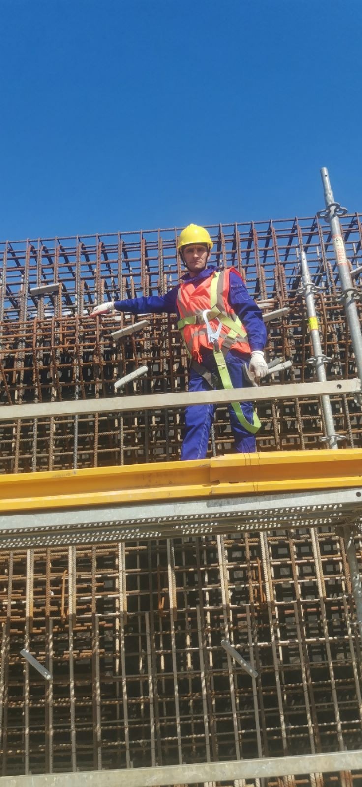 Zbrojarz w kasku i kamizelce na rusztowaniu, wskazujący na siatkę zbrojeniową na tle błękitnego nieba.