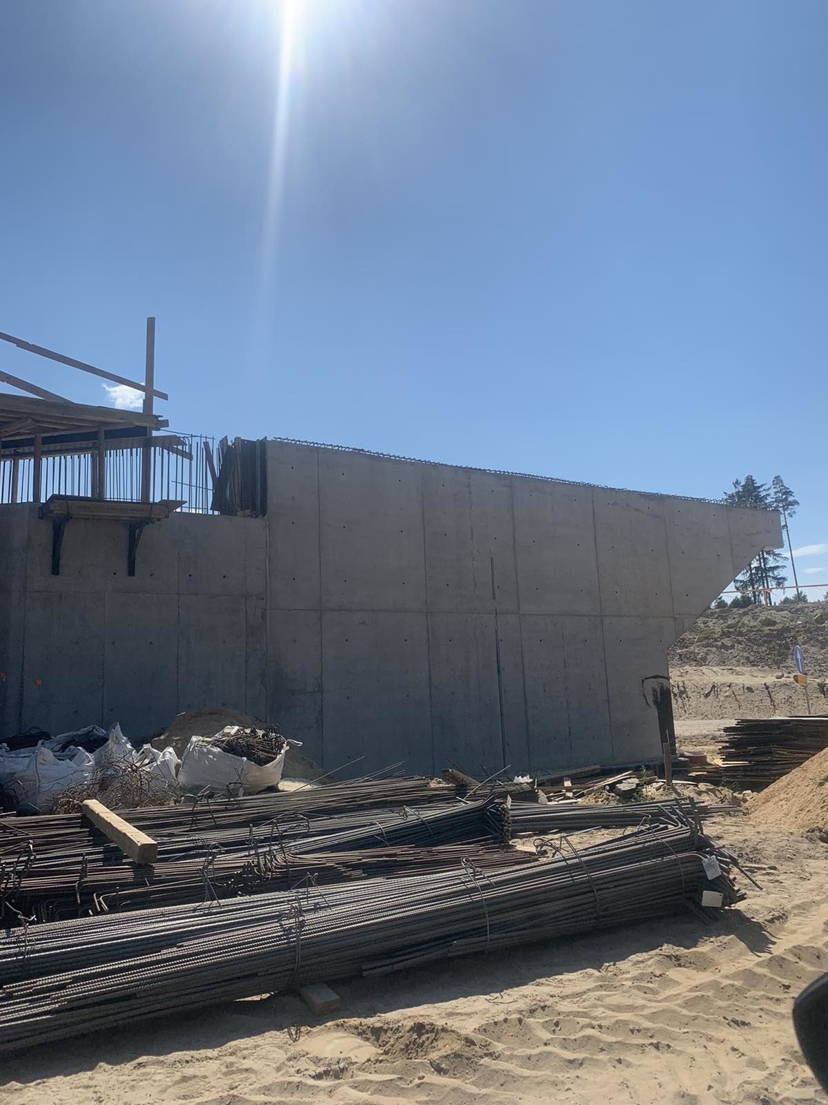 Wznoszenie żelbetowej konstrukcji mostu w surowym stanie, widoczne zbrojenia i szalunki, z hałdą prętów zbrojeniowych na pierwszym planie pod gołym niebem.