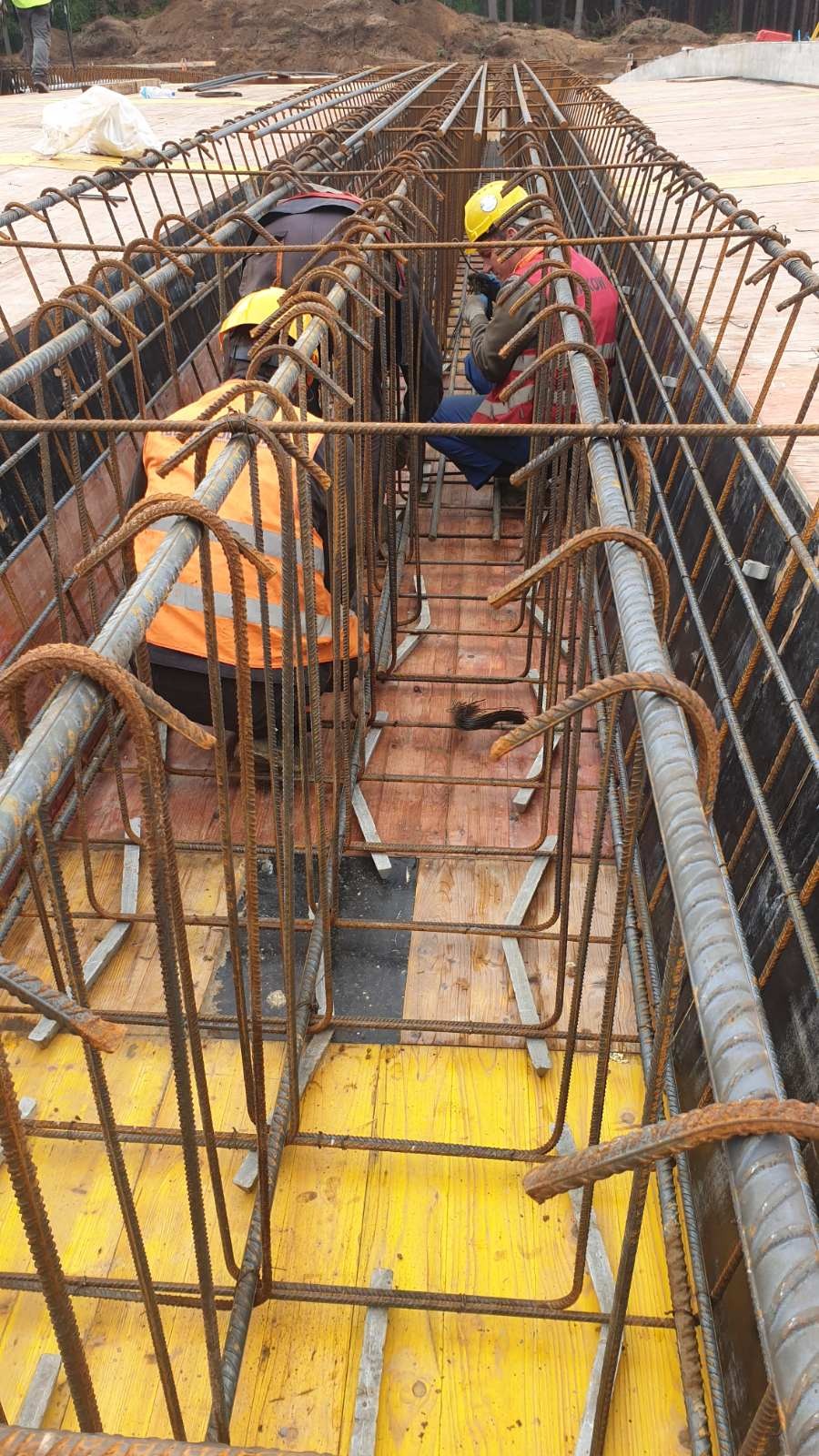Prace zbrojarskie: Widok z góry na szkielet z prętów zbrojeniowych, z pracownikami w kaskach i kamizelkach odblaskowych, przygotowywany na drewnianym szalunku.