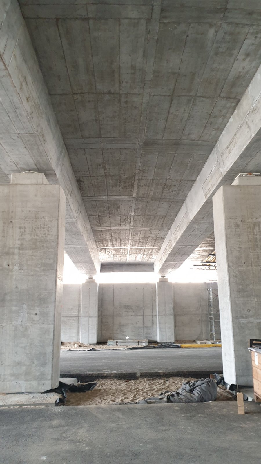 Surowe, betonowe elementy konstrukcyjne mostu w trakcie budowy, widok z dołu ukazujący filary, dźwigary i strop, z piaskiem i materiałami budowlanymi na ziemi.