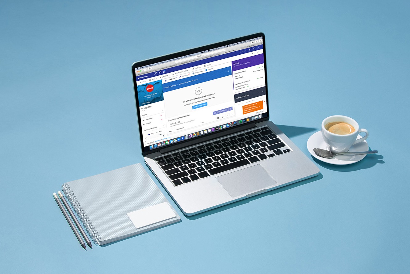 Laptop z otwartym oknem przeglądarki na platformie do zarządzania zadaniami, obok notatnik, wizytówka i filiżanka espresso na jasnoniebieskim tle.