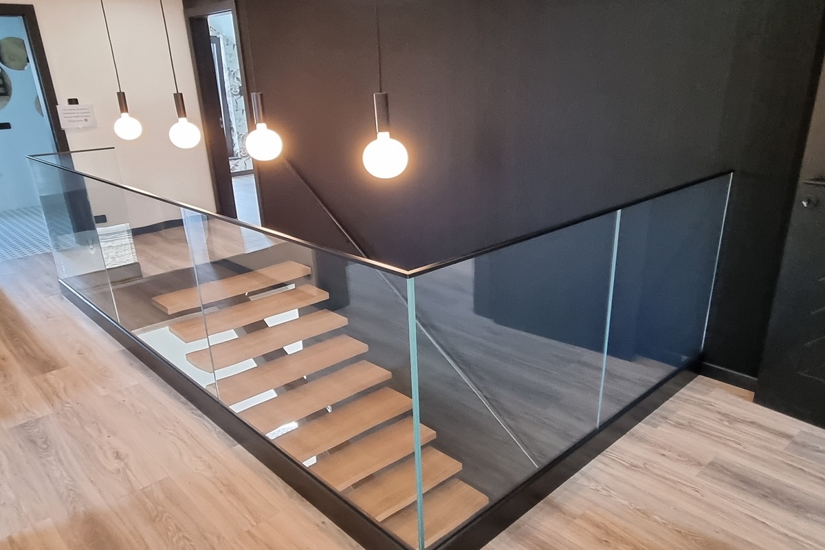 Nowoczesne schody z drewnianymi stopniami zabezpieczone szkłem hartowanym i czarną ramą, oświetlone wiszącymi lampami kulami.