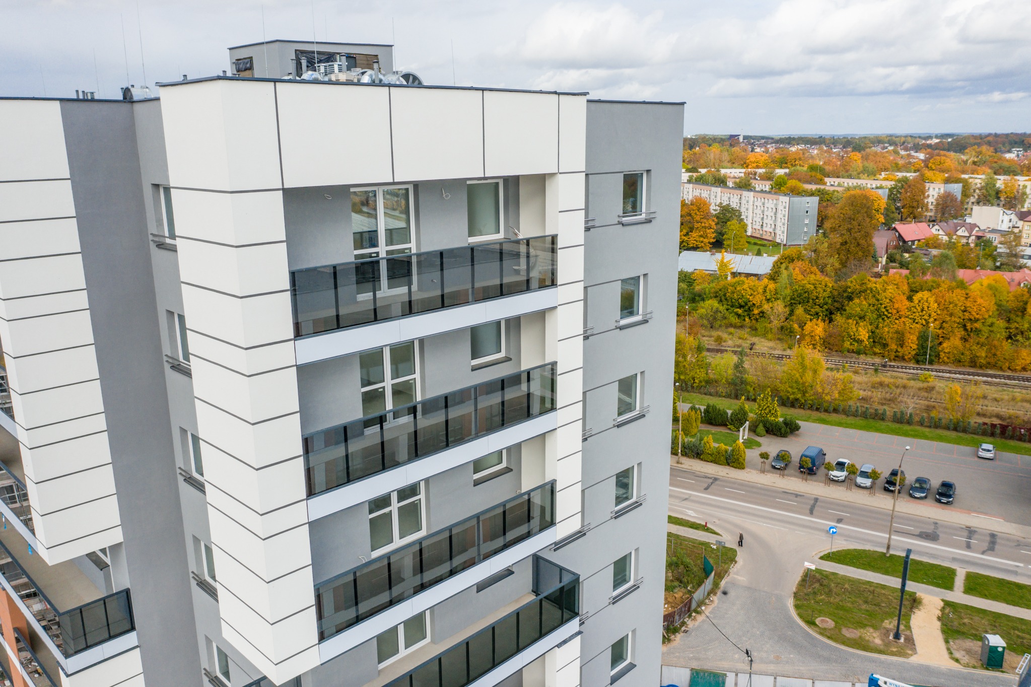 Nowoczesny budynek mieszkalny z balkonami zabezpieczonymi przezroczystymi panelami, widok z góry na tle jesiennego krajobrazu miejskiego.