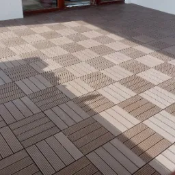 Ułożona podłoga tarasowa z kwadratowych paneli kompozytowych w odcieniach brązu i beżu, ułożonych naprzemiennie, z widocznym cieniem i fragmentem ściany budynku.