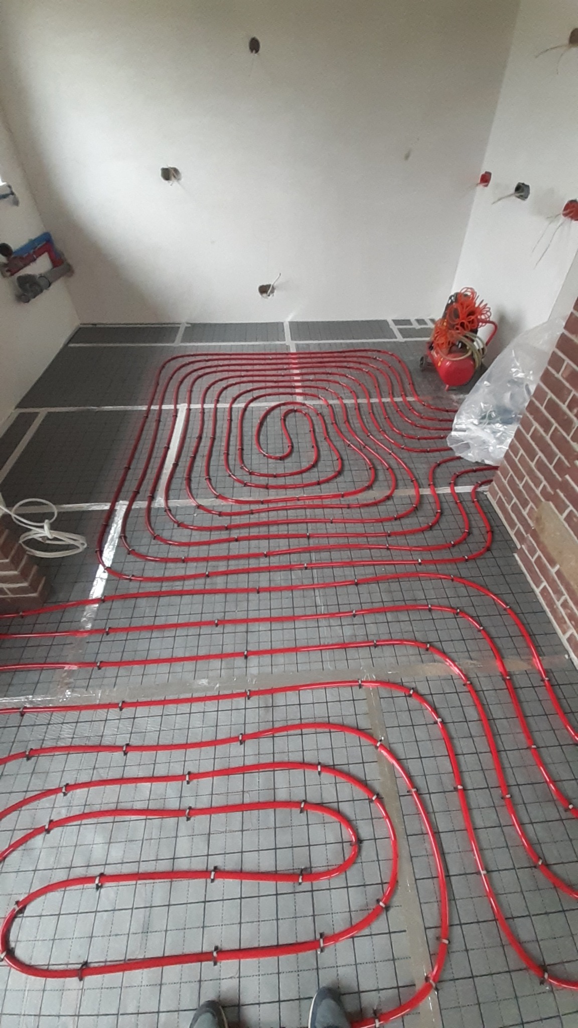 Czerwone rury ogrzewania podłogowego ułożone na siatce w surowym pomieszczeniu z widocznymi instalacjami elektrycznymi i ceglanym fragmentem ściany, obok stoi czerwony kompresor.