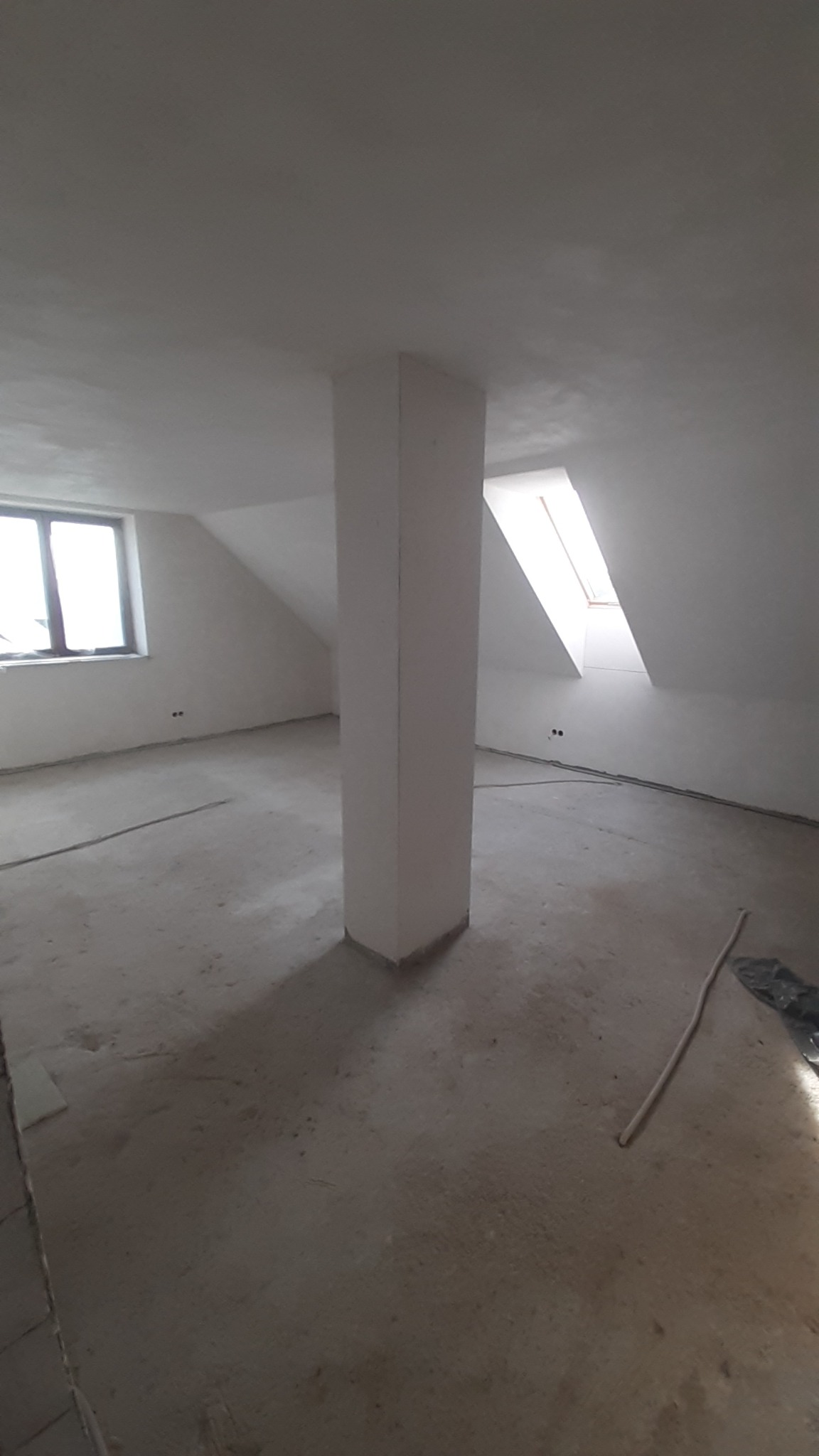 Surowe wnętrze poddasza z kolumną nośną, oknem dachowym i zwykłym oknem, przygotowane do dalszych prac wykończeniowych.