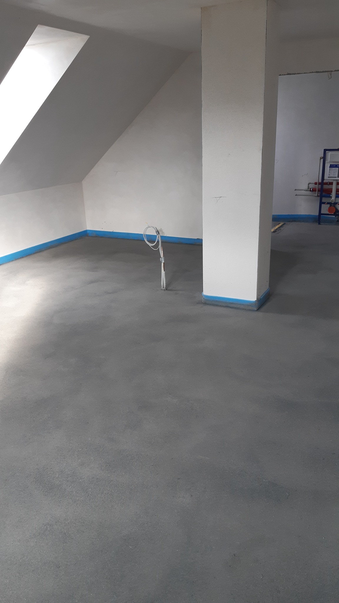 Wylany, szary betonowy podkład podłogowy w pomieszczeniu ze skosami, zabezpieczony niebieską taśmą malarską przy ścianach i filarze, wystające przewody elektryczne.