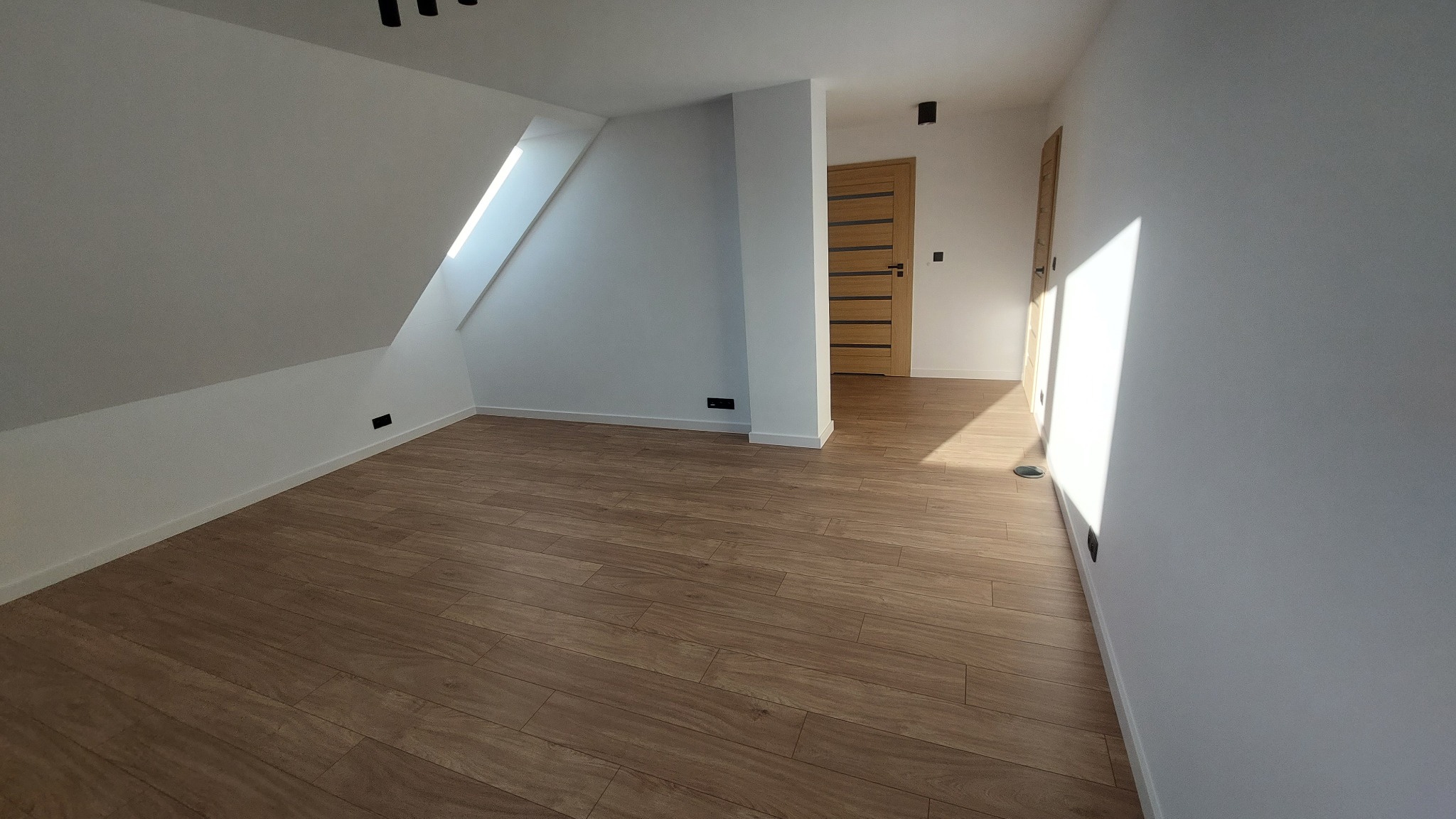 Poddasze po remoncie z nową podłogą z paneli drewnopodobnych, białe ściany, okno dachowe i widoczne drzwi wewnętrzne.