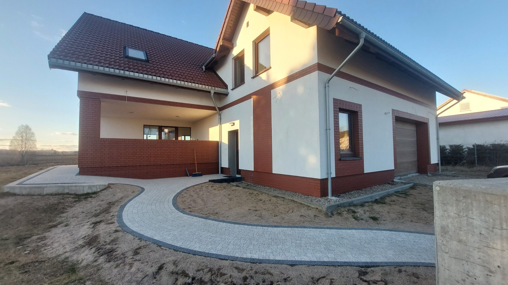 Wykończony dom jednorodzinny z czerwonym dachem i ceglaną elewacją, z brukowanym chodnikiem prowadzącym do wejścia i garażu, sfotografowany w słoneczny dzień.