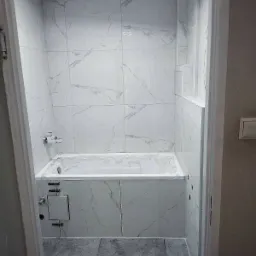 Wykończona łazienka z wanną obłożoną marmurowymi płytkami, widok zza futryny drzwiowej, szara podłoga.