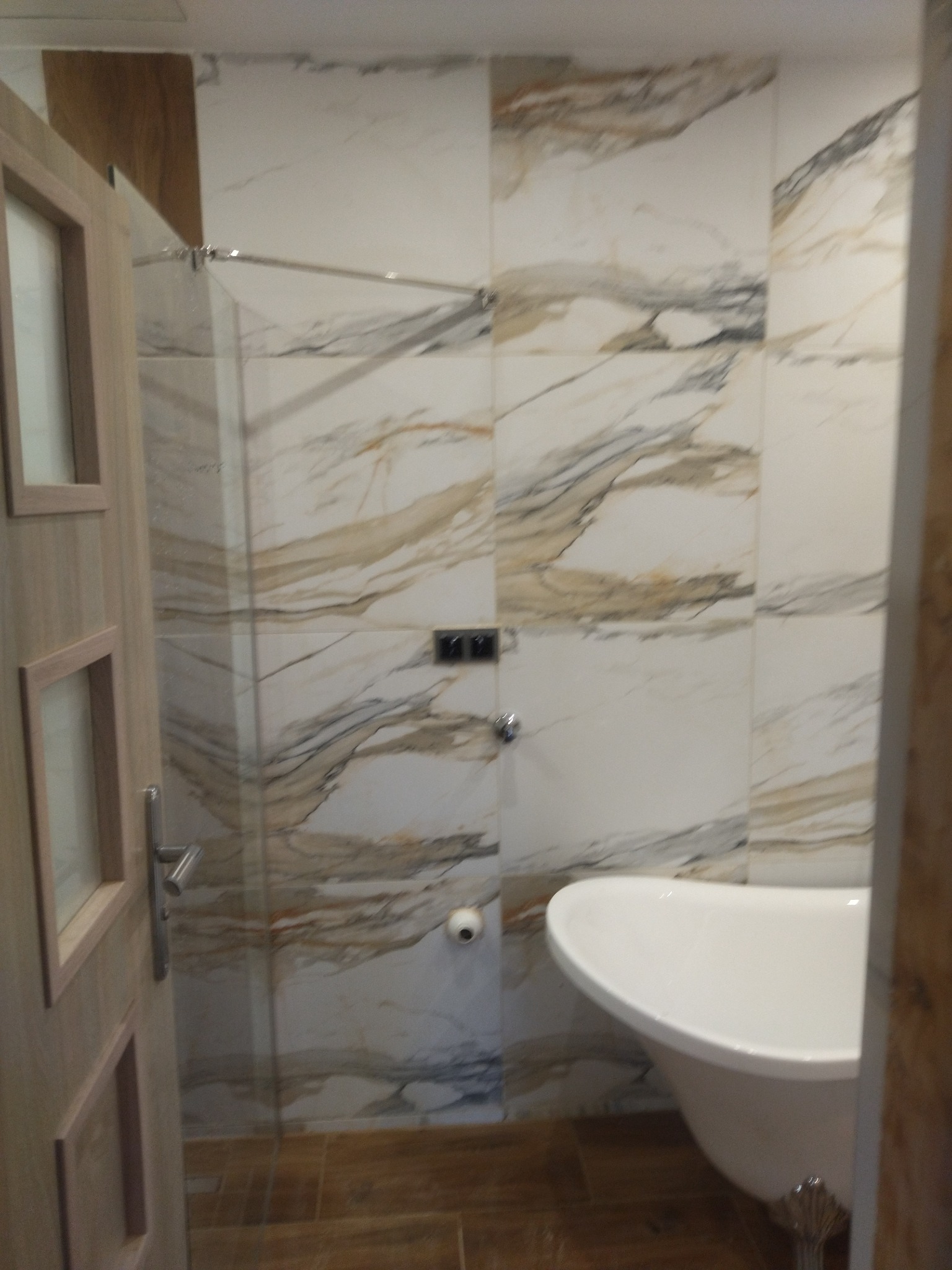 Wykończona łazienka z wanną wolnostojącą na zdobionych nóżkach, ściany wyłożone płytkami imitującymi marmur, widoczny fragment szklanej kabiny prysznicowej i drewniane elementy wykończenia.