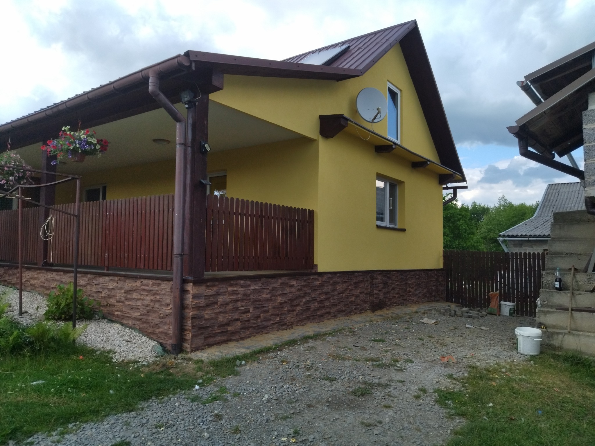 Żółty dom jednorodzinny z brązową elewacją imitującą kamień, brązowym dachem i drewnianym ogrodzeniem werandy, widok z boku na tle pochmurnego nieba, w tle widoczny drugi budynek i rozrzucone...