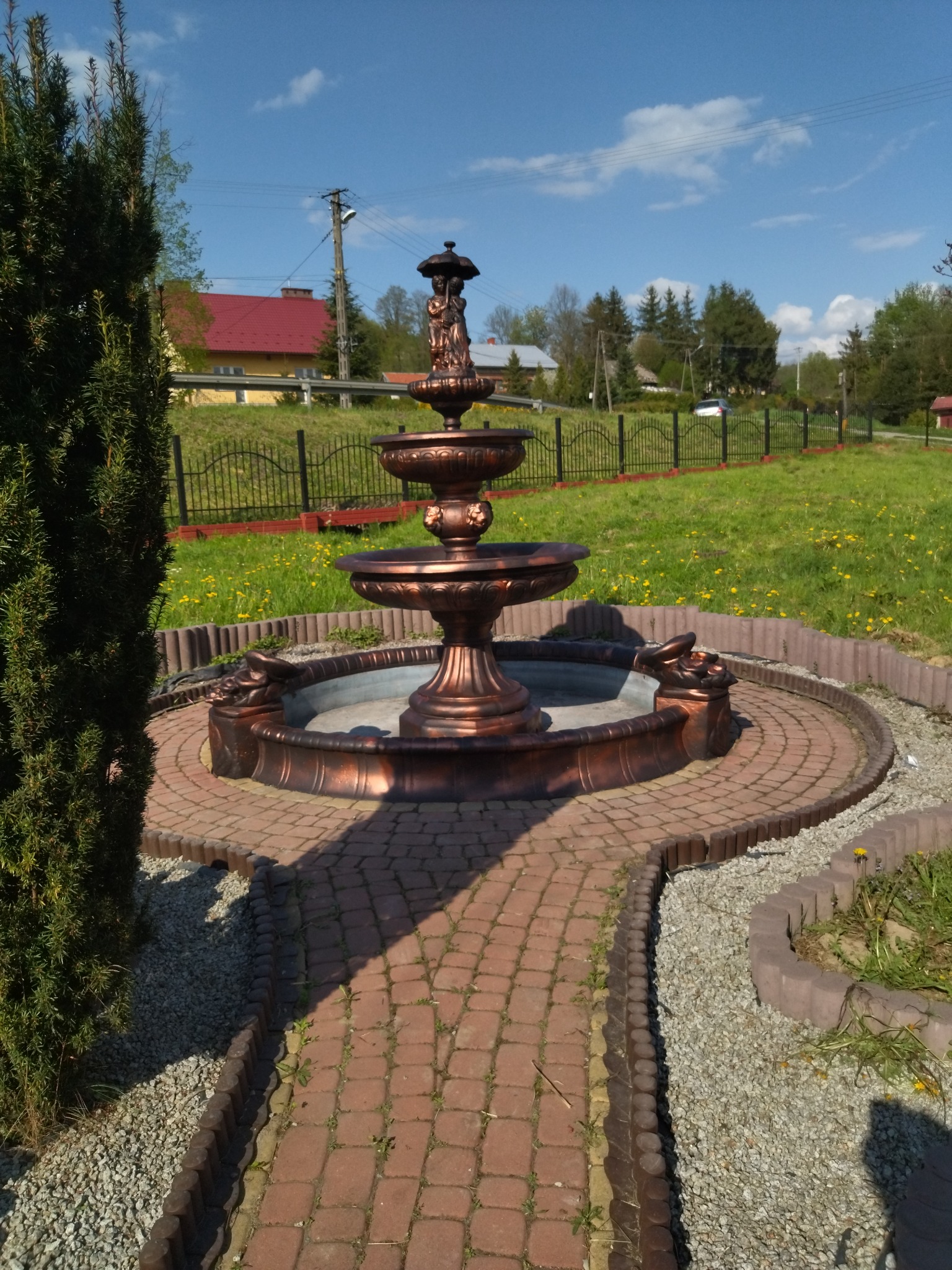 Ozdobna, miedziana fontanna ogrodowa z motywem figur na szczycie, otoczona brukowaną ścieżką i zielenią, na tle ogrodzenia i zabudowań.