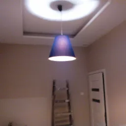 Niebieski klosz lampy wiszącej w pokoju z pomalowanymi ścianami i zamontowaną podwieszaną konstrukcją sufitową, widoczna drabina malarska i częściowo białe drzwi.