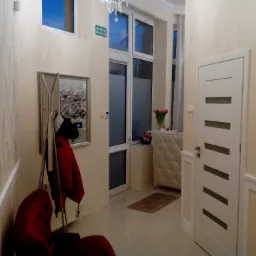 Jasny korytarz z białą, nowoczesną stolarką drzwiową z poziomymi przeszkleniami, w tle widoczne okno, wieszak na ubrania z czerwoną kurtką i dekoracyjny żyrandol.