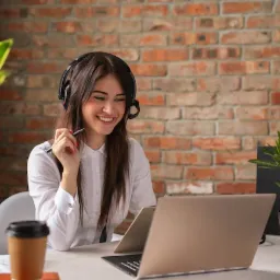 Uśmiechnięta kobieta z długimi brązowymi włosami w słuchawkach z mikrofonem, siedząca przy biurku z laptopem, kubkiem kawy i roślinami, na tle ściany z czerwonej cegły.