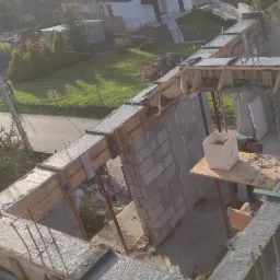 Wylewanie betonu w szalunkach na budowie domu jednorodzinnego, widok z g&oacute;ry na konstrukcję zbrojoną, w tle zabudowa mieszkalna i zalesione wzg&oacute;rza.