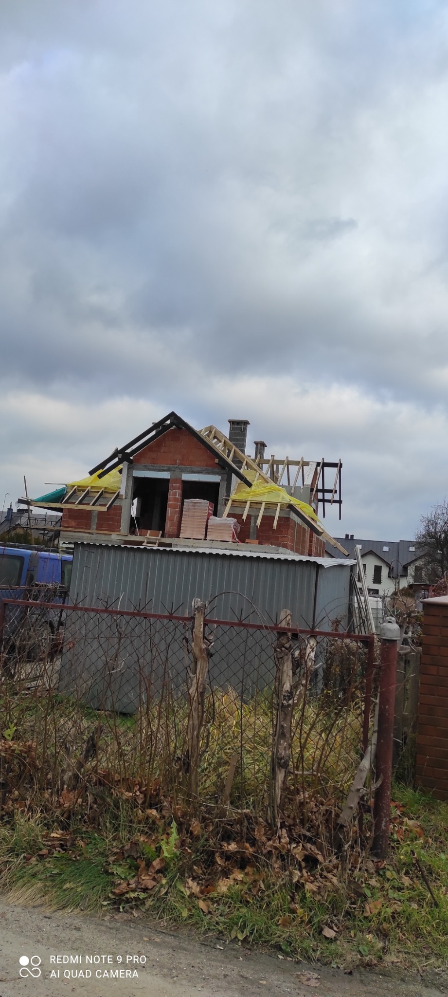 Surowy stan domu w trakcie budowy z widoczną konstrukcją dachu, cegłami i elementami drewnianymi, sfotografowany zza ogrodzenia w pochmurny dzień.