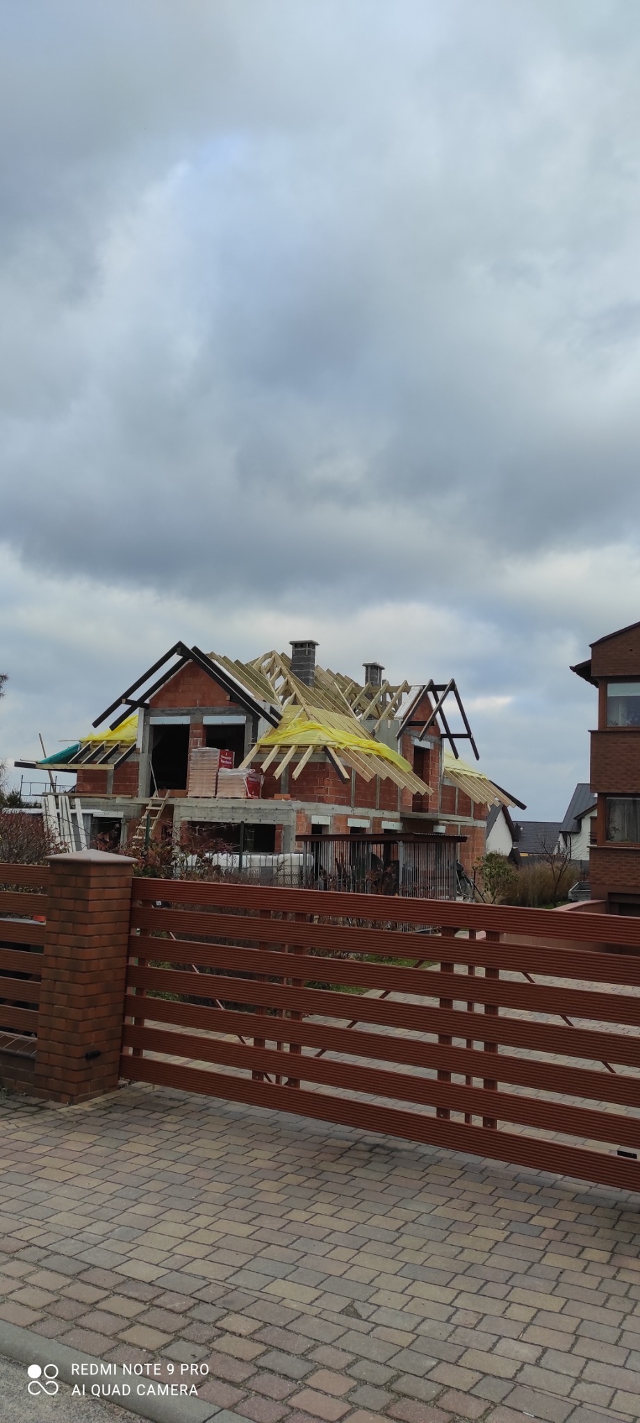 Budowa domu murowanego w stanie surowym otwartym z widoczną konstrukcją dachu, ceglanymi ścianami i oknami bez szyb, widziana zza brązowego ogrodzenia z ceglanym słupkiem.