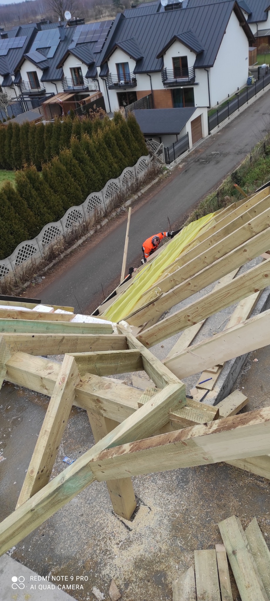 Konstrukcja drewniana dachu w trakcie budowy domu w Toporzysko, widoczny pracownik w pomarańczowej kurtce oraz fragment osiedla w tle.