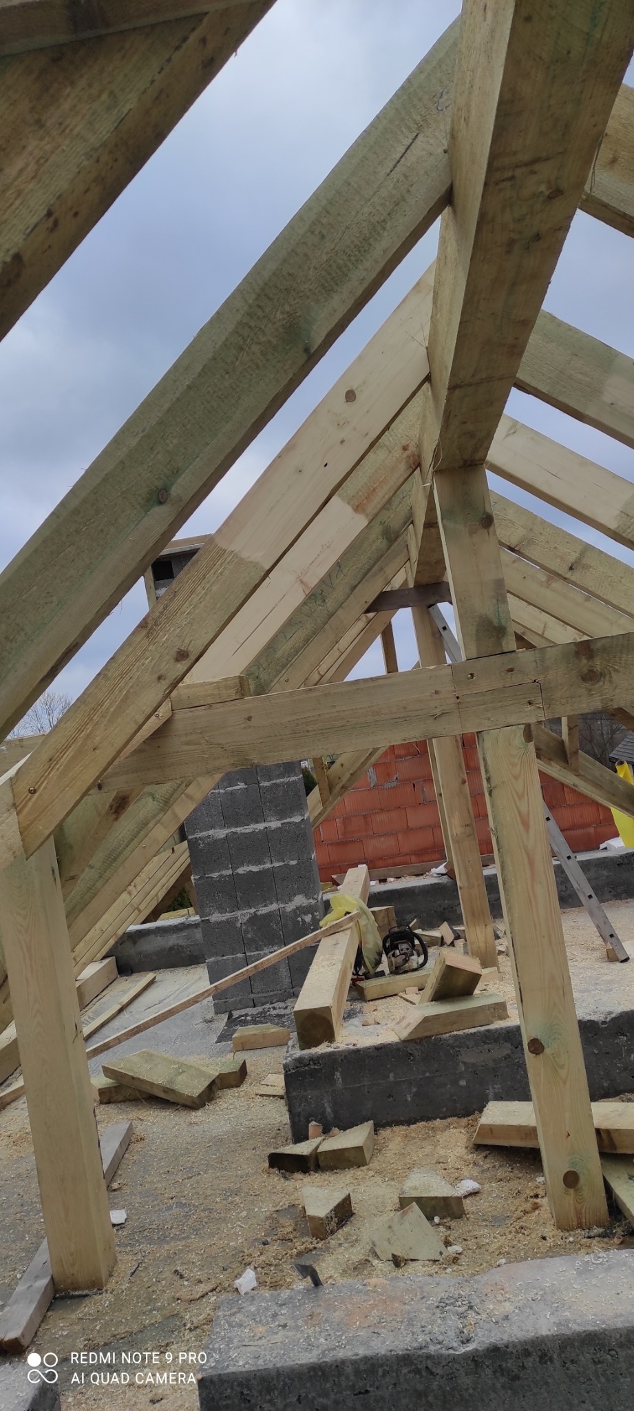 Konstrukcja drewniana dachu w trakcie budowy, widoczne krokwie, mur z cegły i pustaków, oraz piła łańcuchowa i drabina oparta o ścianę.