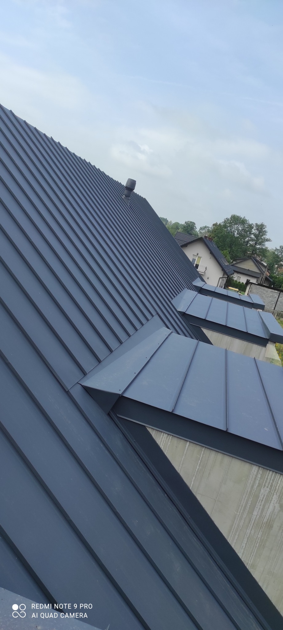 Nowoczesny dach z blachy na rąbek stojący w kolorze grafitowym, z widocznymi lukarnami i kominem wentylacyjnym, na tle pochmurnego nieba i zabudowy jednorodzinnej.