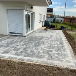 Nowo ułożony taras z szarej kostki brukowej z jasną obwódką przy białym budynku z szarą roletą, widoczny wózek i ubijarka w tle