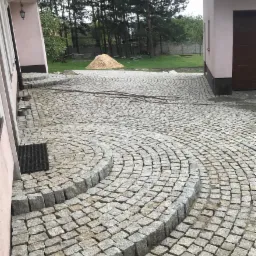 Granitowa kostka brukowa ułożona w łukowaty wzór z niskimi schodkami, widoczny wąż ogrodowy, w tle budynek i stos piasku.