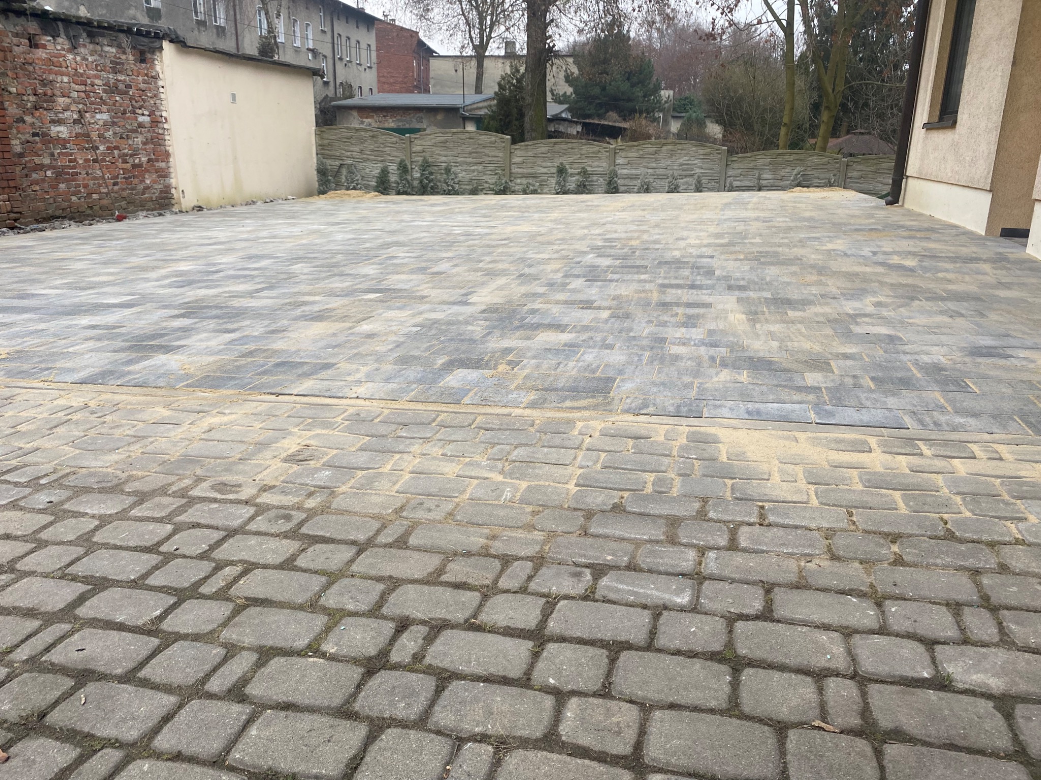 Nowo ułożona, szara kostka brukowa na dużej powierzchni podwórza, z widocznym piaskiem pomiędzy kostkami, obok mur z cegły i budynki w tle.