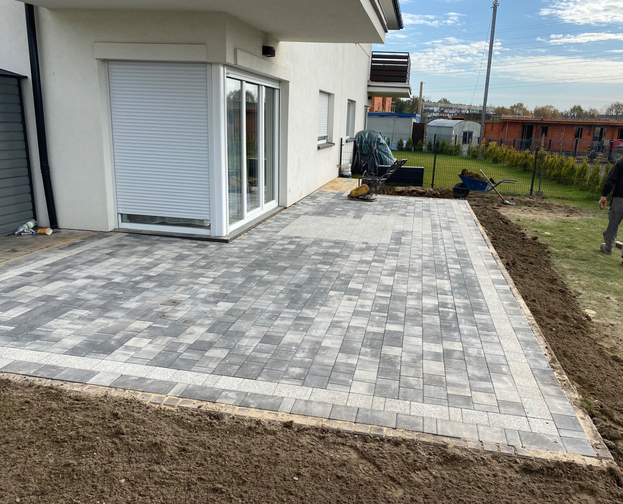 Nowo ułożony taras z szarej kostki brukowej z jasną obwódką przy białym budynku z szarą roletą, widoczny wózek i ubijarka w tle