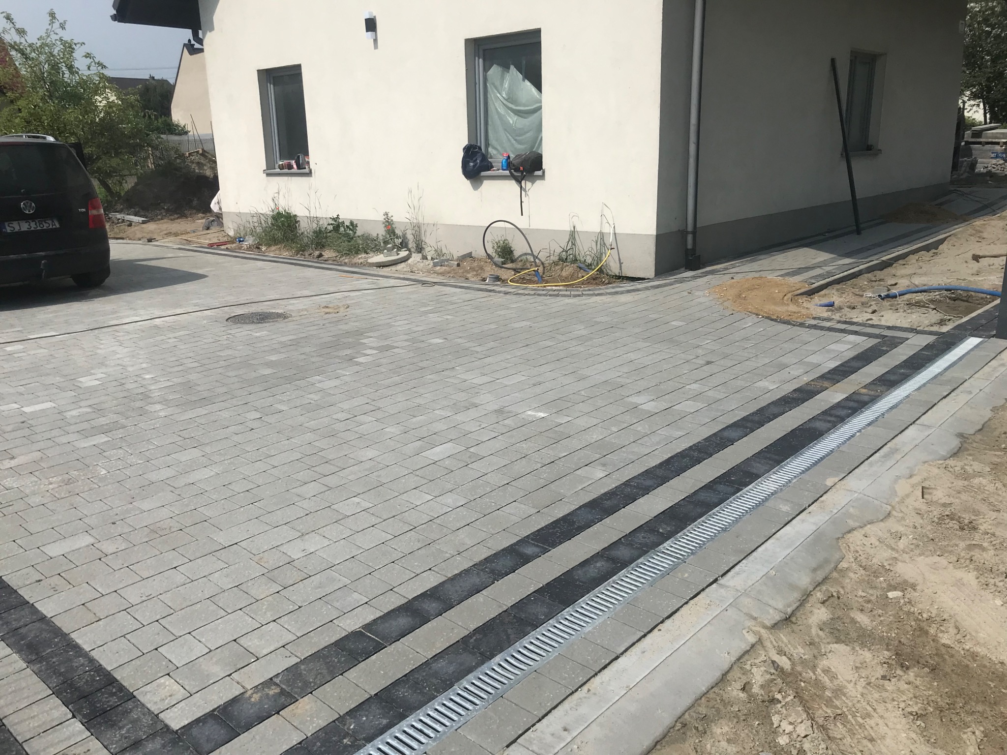 Nowo ułożona szara kostka brukowa z ciemnymi pasami i odpływem liniowym przed białym domem z oknem, częściowo zasłoniętym folią.