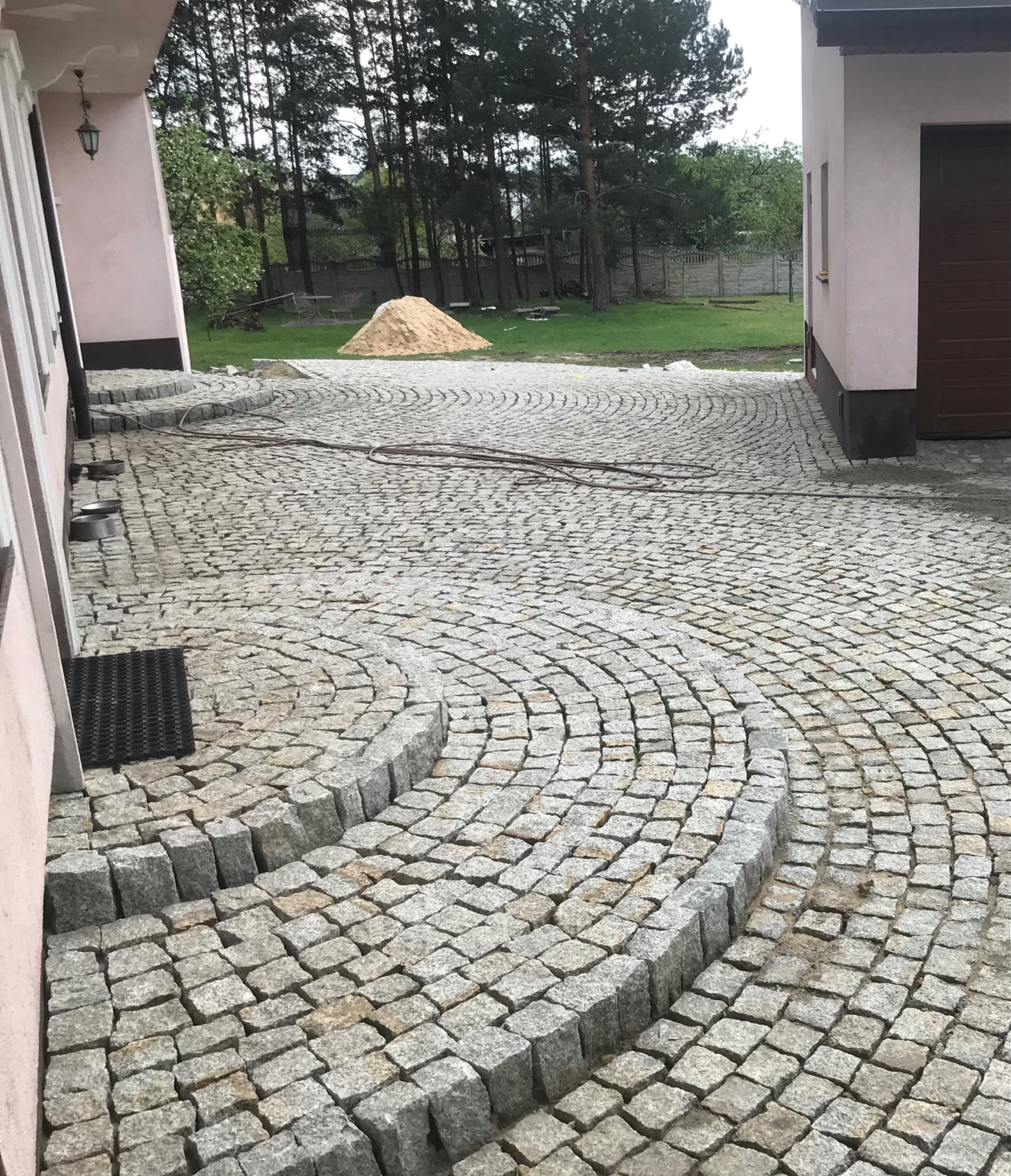 Granitowa kostka brukowa ułożona w łukowaty wzór z niskimi schodkami, widoczny wąż ogrodowy, w tle budynek i stos piasku.