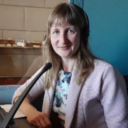 Uśmiechnięta tłumaczka w kabinie z mikrofonem i słuchawkami, w tle bufet z przekąskami za szybą.