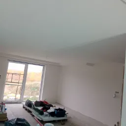 Wnętrze pokoju w trakcie prac wykończeniowych, świeżo pomalowane ściany i sufit, widoczne narzędzia i materiały budowlane na podłodze, okno z widokiem na zewnątrz.