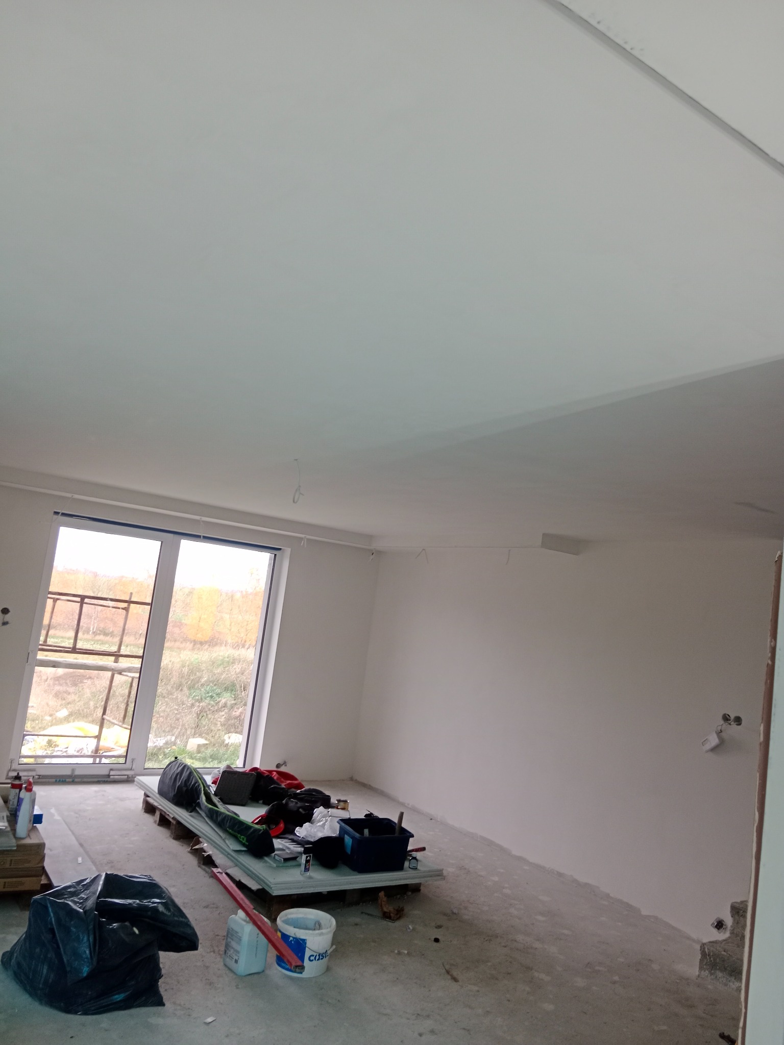 Wnętrze pokoju w trakcie prac wykończeniowych, widoczne gładkie, białe ściany i sufit, okno z widokiem na zewnątrz, podłoga przygotowana do dalszych prac, narzędzia i materiały budowlane...