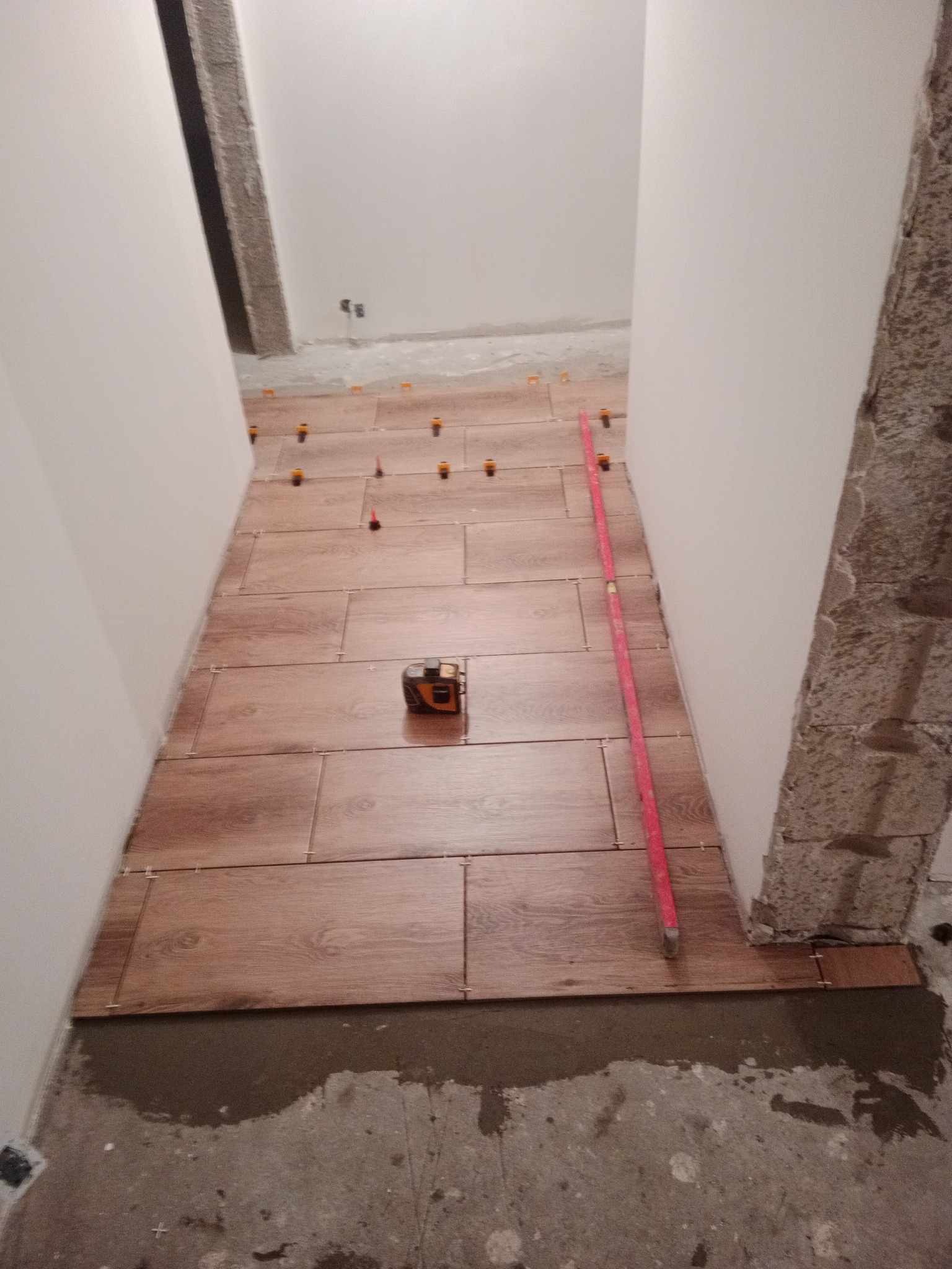 Układanie podłogi z płytek imitujących drewno w wąskim korytarzu, widoczne poziomica, miara laserowa i system poziomowania płytek.