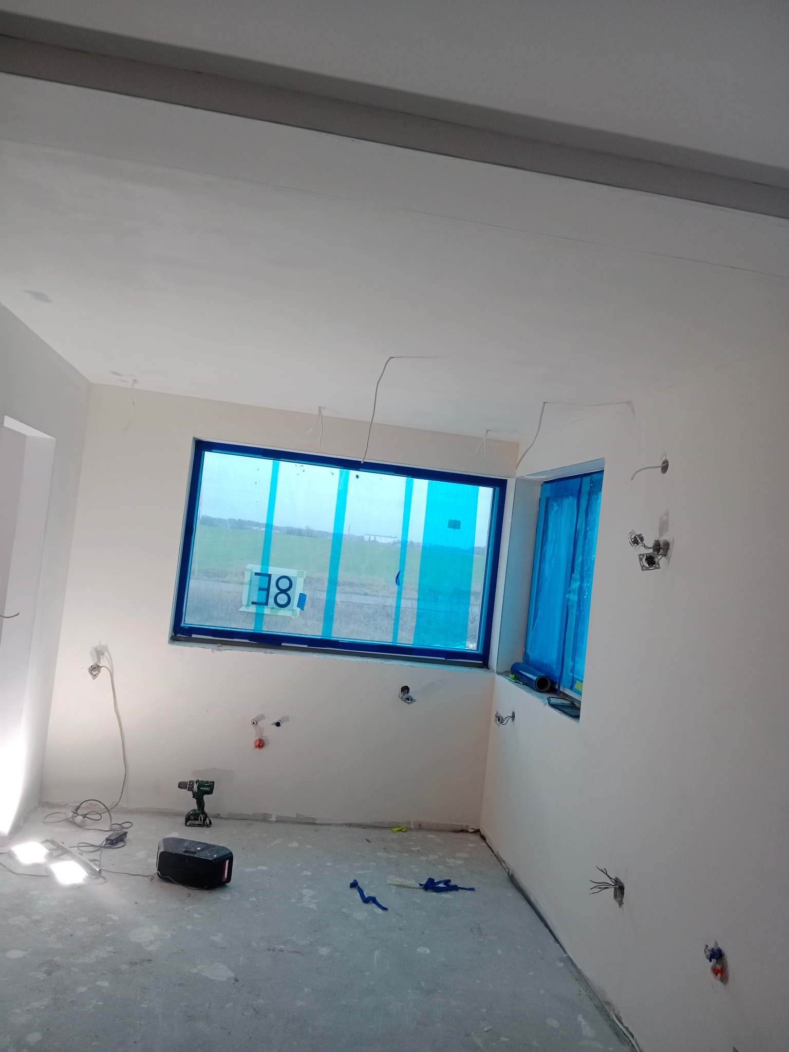 Wnętrze w trakcie prac przygotowawczych: zabezpieczone okna folią, świeżo położona gładź, widoczne puszki elektryczne, narzędzia (wiertarka) i lampa na podłodze.