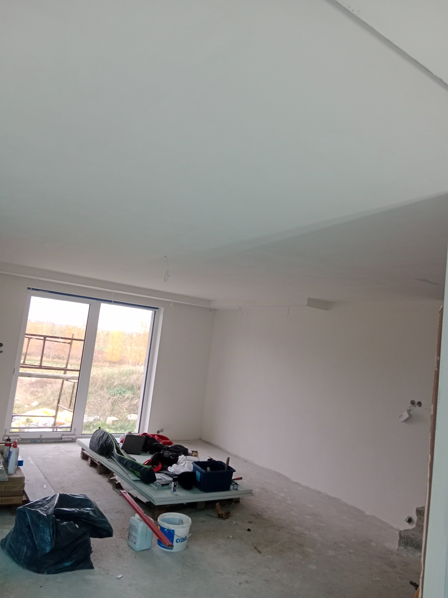 Wnętrze pokoju w trakcie prac wykończeniowych, świeżo pomalowane ściany i sufit, widoczne narzędzia i materiały budowlane na podłodze, okno z widokiem na zewnątrz.
