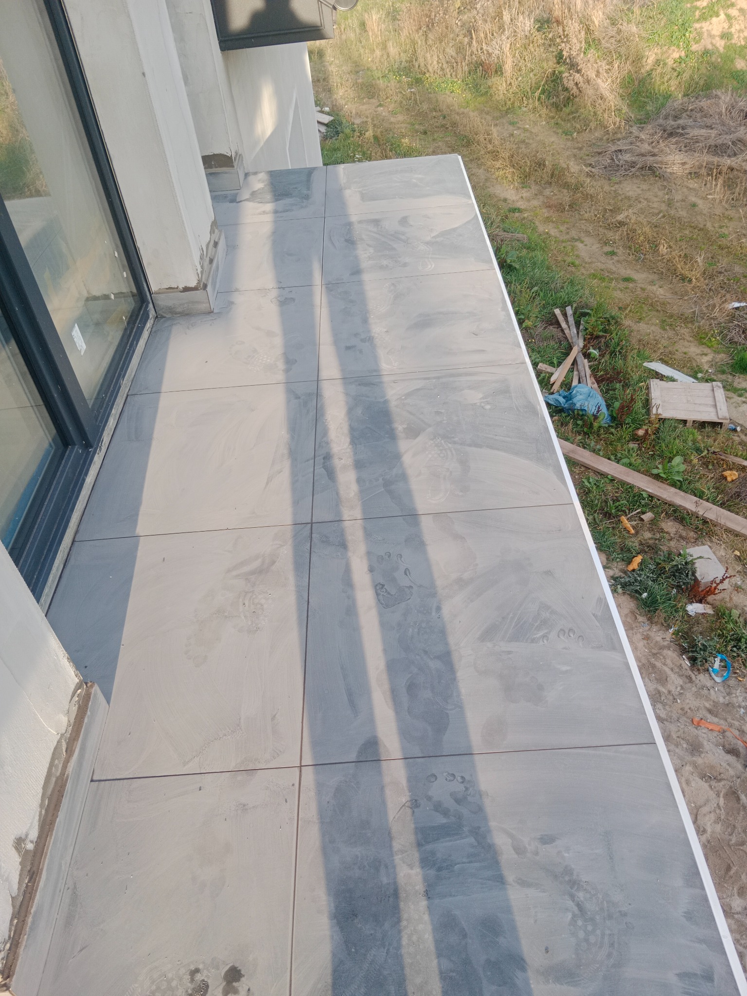 Szare płytki balkonowe ułożone na zewnątrz, widoczne ślady stóp i częściowo zabrudzenia po montażu, otoczenie w stanie surowym.