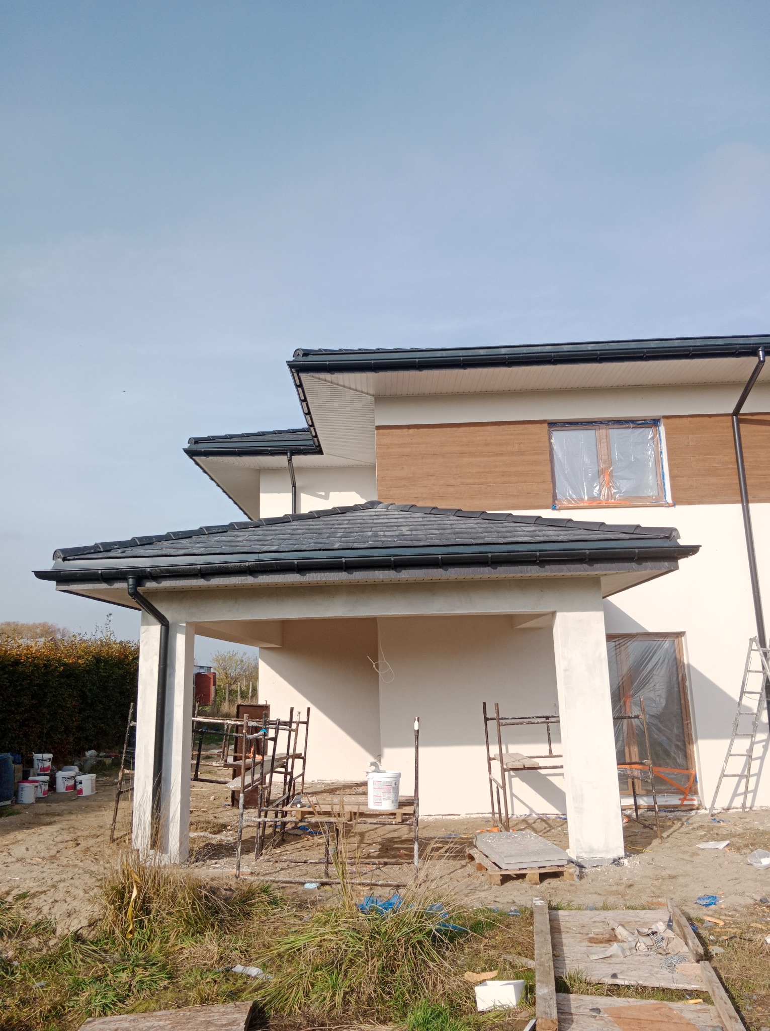 Nowoczesny dom w trakcie prac elewacyjnych: zabezpieczone okna folią, widoczne rusztowania i puszki z farbą na zewnątrz.