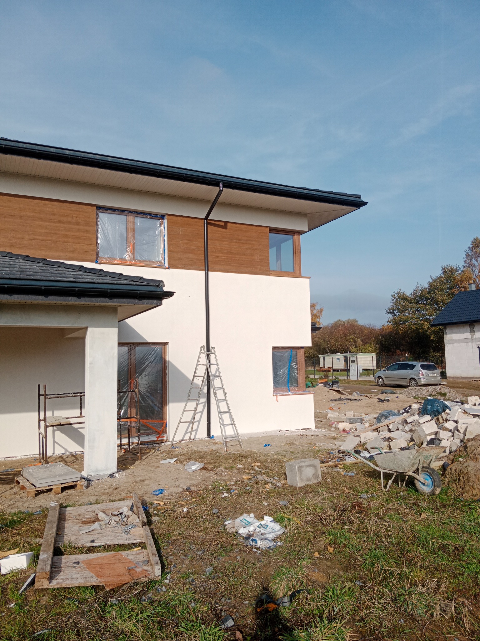 Dwukondygnacyjny dom z oknami zabezpieczonymi folią, elewacja częściowo otynkowana na biało, część wykończona drewnianą boazerią, na trawniku gruz i taczka, drabina oparta o ścianę.