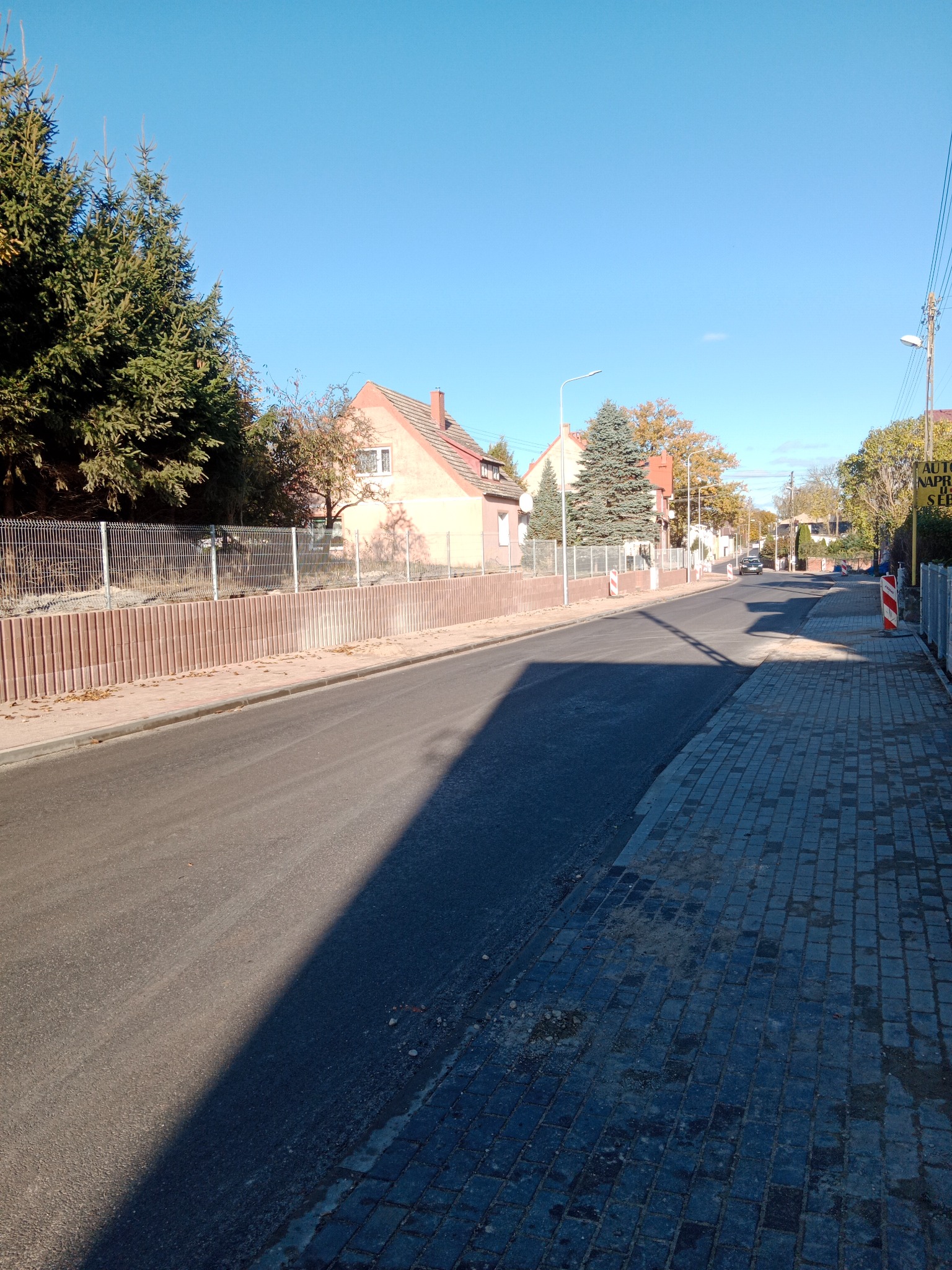 Ulica w małym miasteczku z nową nawierzchnią asfaltową i chodnikiem z kostki brukowej, w tle domy jednorodzinne i zieleń.