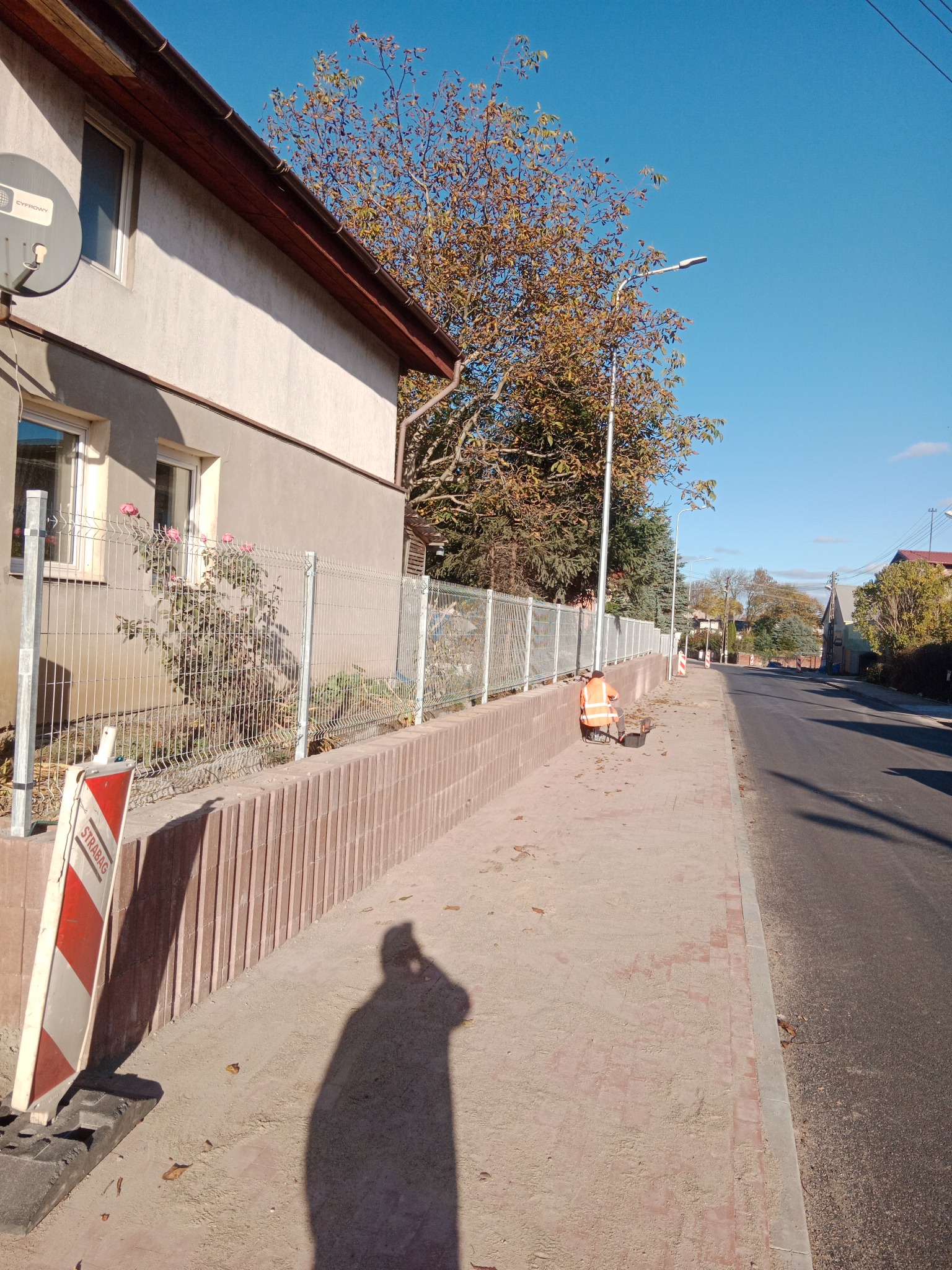 Pracownik w pomarańczowej kamizelce czyszczący chodnik z piasku i liści przy użyciu dmuchawy, po ułożeniu kostki brukowej wzdłuż ogrodzenia posesji.