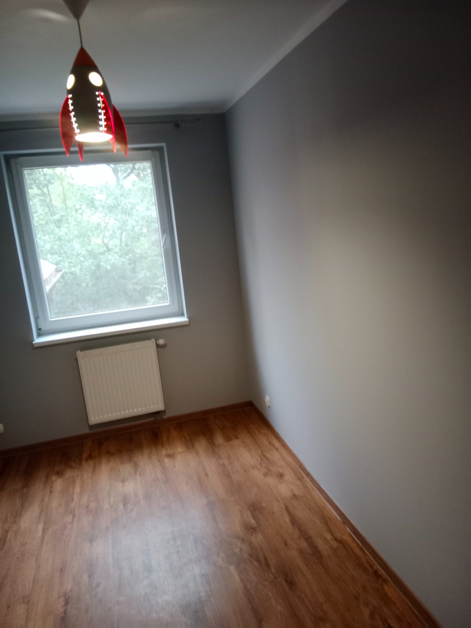 Pomalowane na szaro wnętrze pokoju z drewnianą podłogą, białym grzejnikiem pod oknem i lampą w kształcie rakiety.