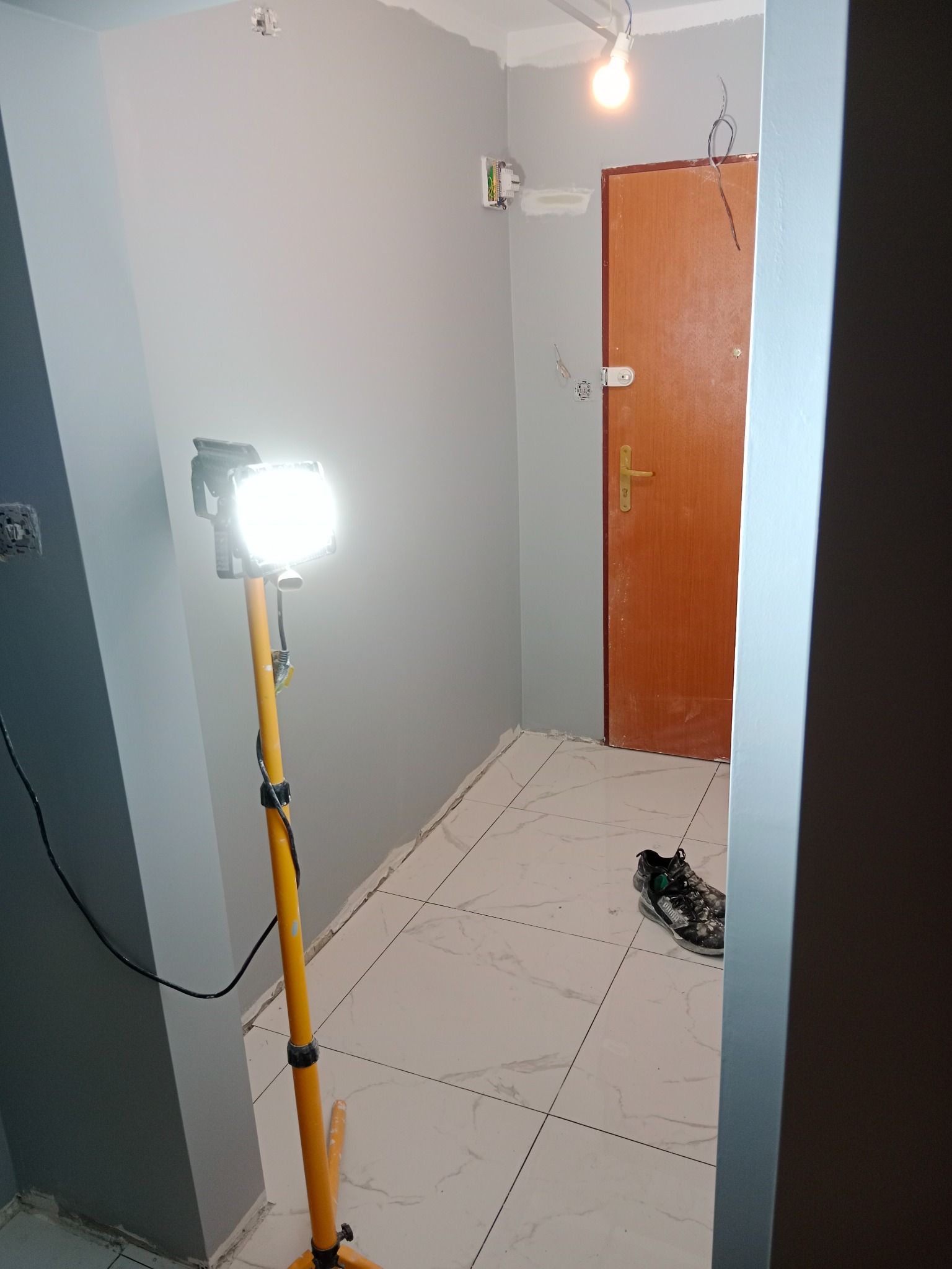Świeżo pomalowane szare ściany w korytarzu, widoczna lampa budowlana na statywie, białe płytki podłogowe i para butów sportowych przy drzwiach.