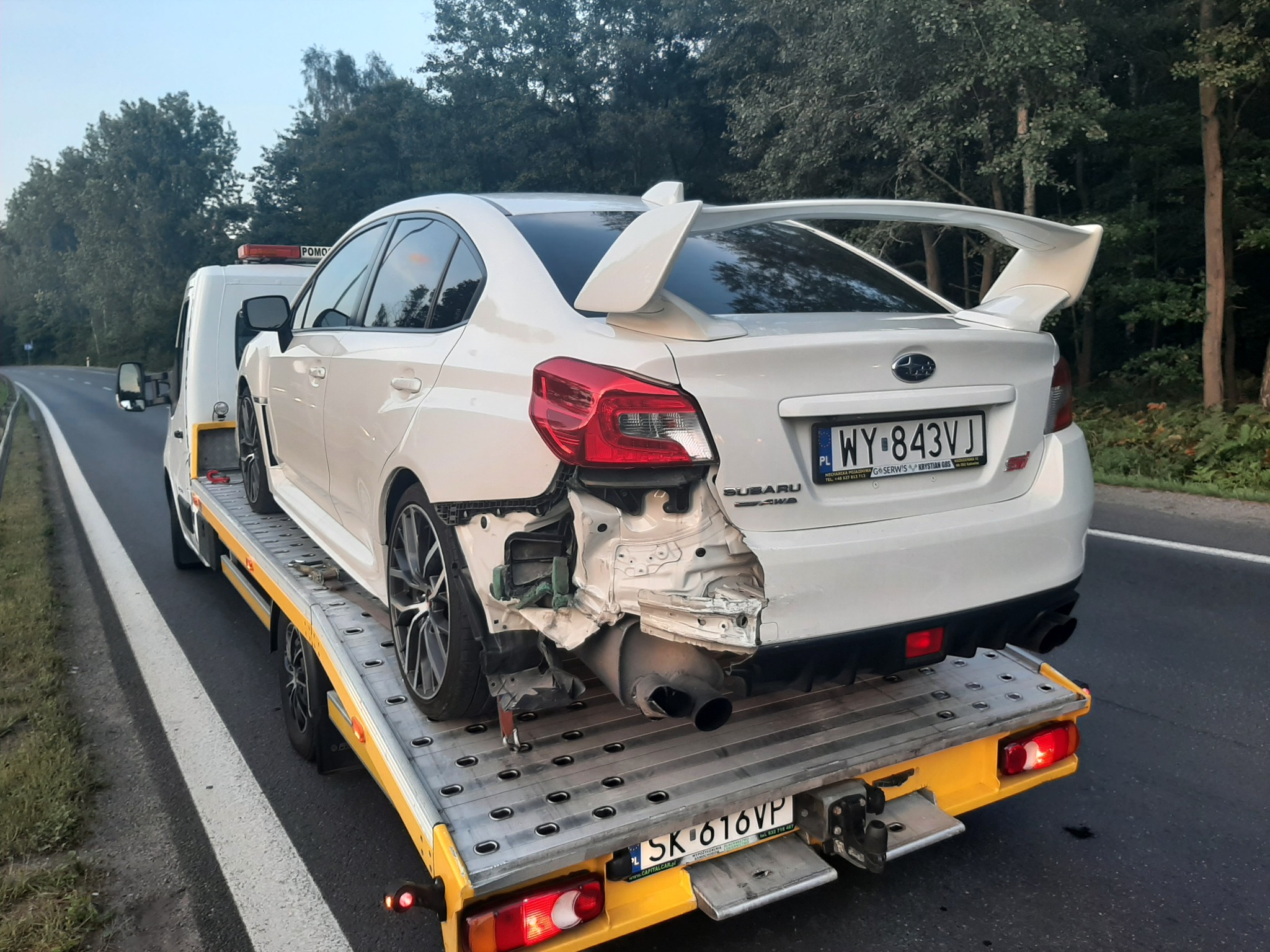 Uszkodzony biały samochód Subaru z dużym spojlerem, z widocznymi uszkodzeniami tylnej części nadwozia, transportowany na żółtej lawecie. Widoczna tablica rejestracyjna z Polski.