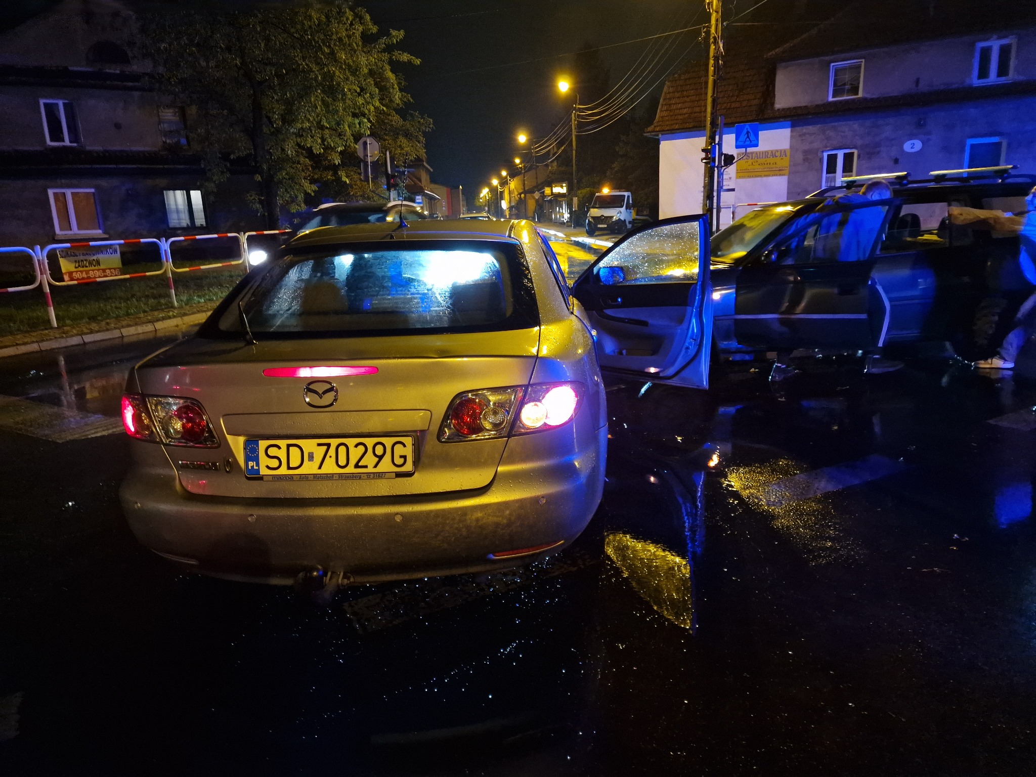 Uszkodzony srebrny samochód Mazda z otwartymi drzwiami na mokrej ulicy nocą, widoczne światła uliczne i budynki w tle, tablica rejestracyjna SD 7029G.