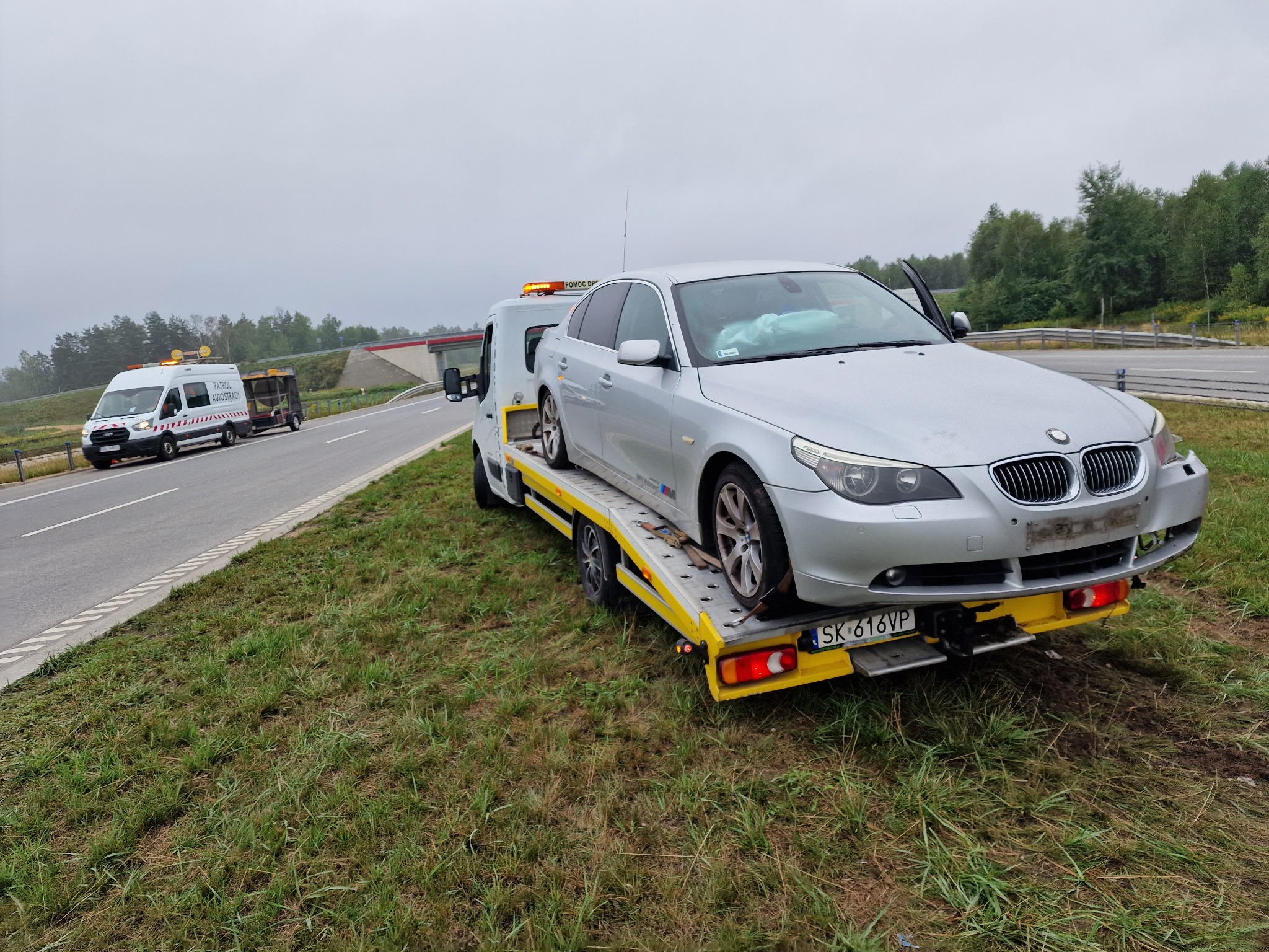 Srebrny samochód osobowy BMW z otwartymi drzwiami kierowcy, przewożony na żółtej lawecie na poboczu drogi, w tle widoczny pojazd pomocy drogowej.