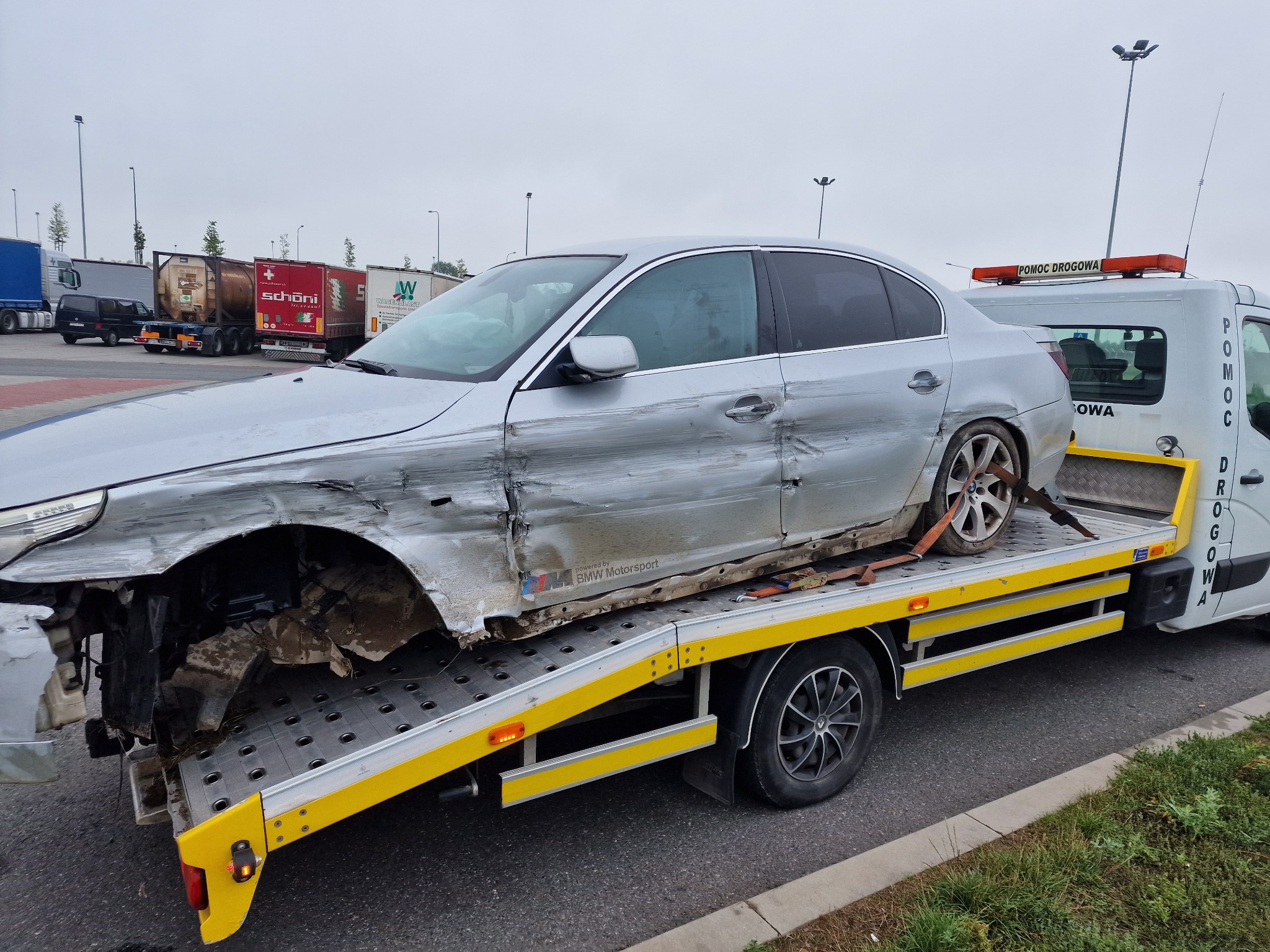 Uszkodzony srebrny samochód BMW Motorsport załadowany na lawetę pomocy drogowej na parkingu, widoczne ciężarówki w tle.